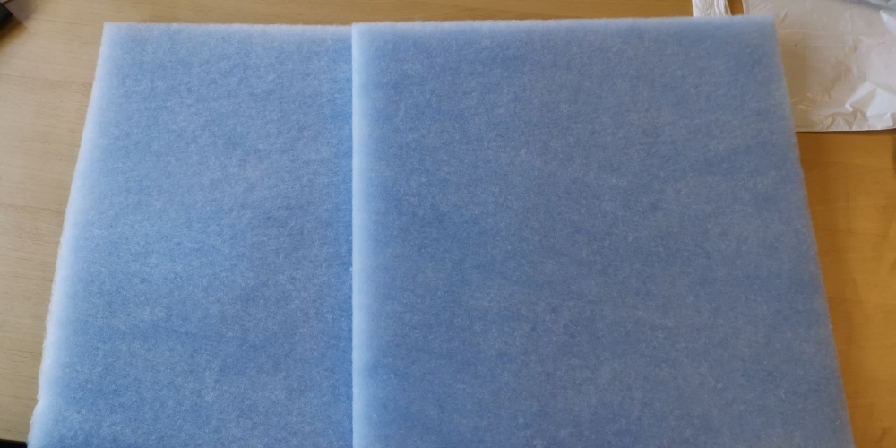 Et rektangulært, lyseblåt filter foldet på midten, med en synlig tekstur, der indikerer dets funktion som et filtreringsmateriale. I øverste højre hjørne er en lille del af et stykke papir synligt.